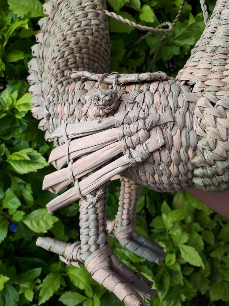 May include: A woven straw rooster figurine in shades of brown and tan. The rooster has a long tail and a detailed head with a comb and wattle. The rooster is standing on two legs with three toes each.