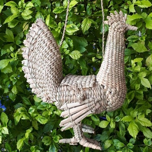 May include: A woven straw rooster-shaped handbag with a rope handle. The handbag is hanging from a tree branch.