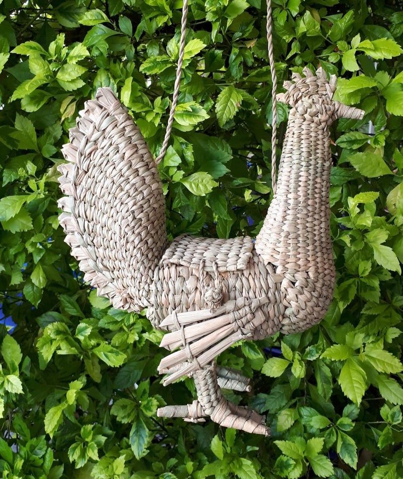 May include: A woven straw rooster-shaped handbag with a rope handle. The handbag is hanging from a tree branch.