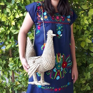May include: A woman wearing a blue embroidered dress with colourful flowers holds a woven straw chicken-shaped bag.