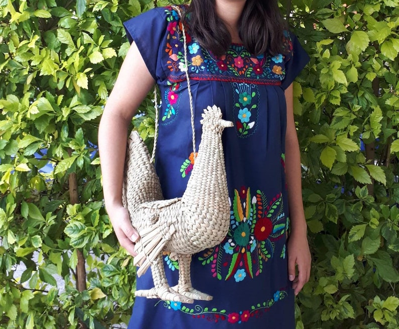 May include: A woman wearing a blue embroidered dress with colourful flowers holds a woven straw chicken-shaped bag.