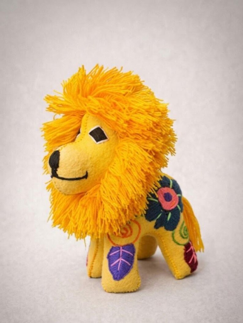 May include: A yellow plush lion toy featuring a thick, bright yellow yarn mane, a black stitched nose and mouth, and diamond-shaped eyes. The body is decorated with colorful embroidered floral and leaf patterns in blue, pink, purple, and green.