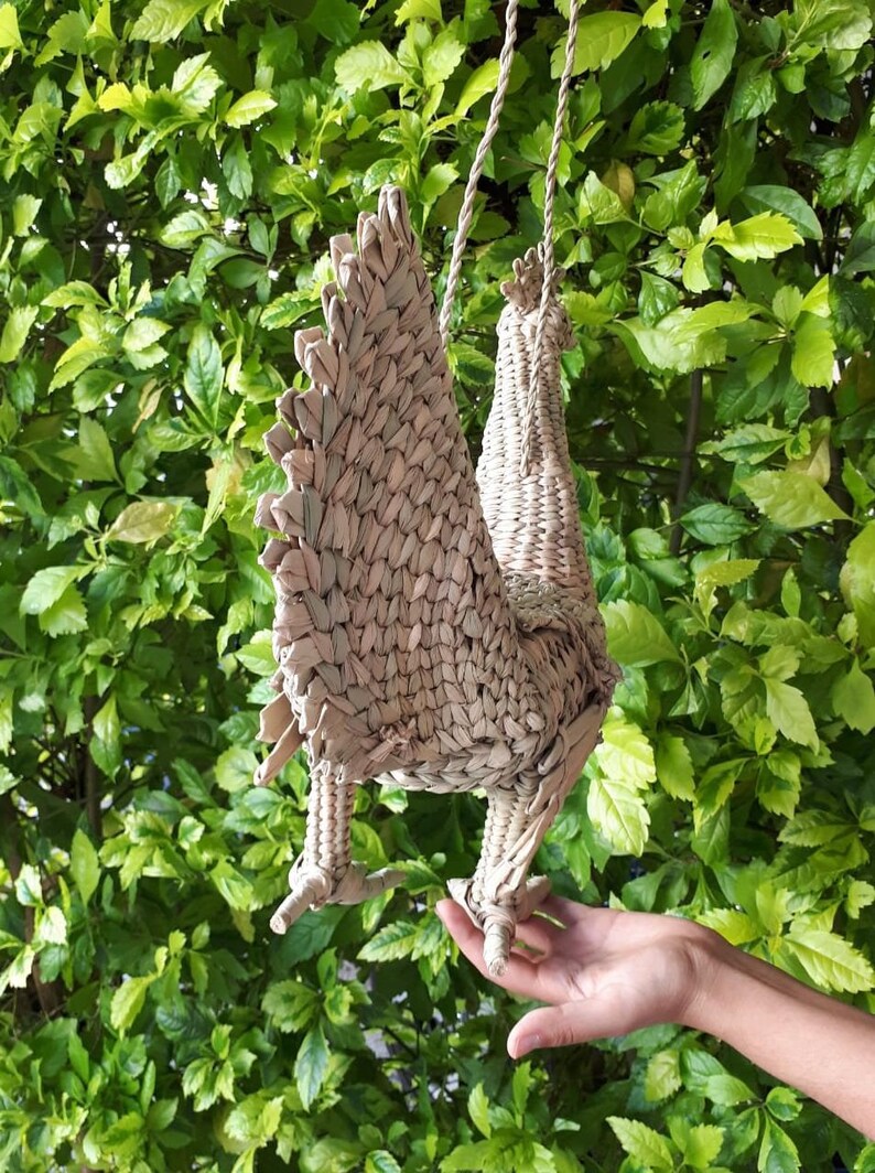 May include: A woven wicker rooster figurine with a long tail, hanging from a rope. The rooster is light brown in colour and has a detailed design.