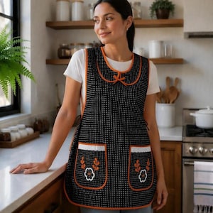 May include: A black apron with a white polka dot pattern, featuring orange trim along the edges and neckline. The apron includes two front pockets, each decorated with white lace trim and an embroidered orange and white floral design.