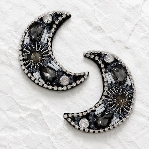 May include: Two crescent moon-shaped patches featuring black felt bases adorned with clear, grey, and black crystals, white seed beads, and silver-toned metal accents. The edges are lined with a row of small, clear, square-cut rhinestones.