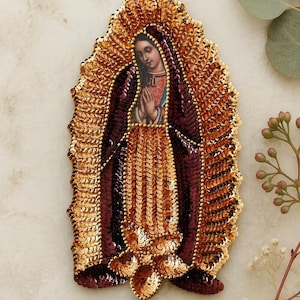 May include: An embroidered patch featuring the Virgin of Guadalupe, adorned with metallic gold and deep burgundy sequins. The central figure is depicted with hands clasped in prayer, surrounded by a radiant, sequined gold halo and dark robe.