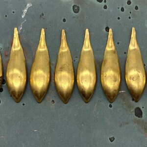 May include: Eight gold-colored, teardrop-shaped metal objects arranged in a row. Each piece has a pointed top and a rounded base. The objects are on a mottled, greenish-gray surface.