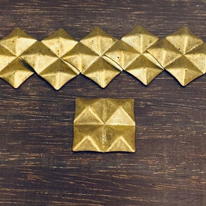 May include: A row of eight gold-colored pyramid-shaped metal embellishments on a dark brown wooden surface. A single embellishment is centered below the row.