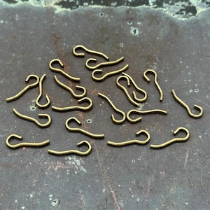 May include: A collection of small, brass-colored metal clasps scattered on a textured, dark surface. Each clasp has a curved hook shape, ideal for jewelry making or craft projects.