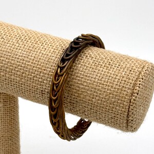 May include: A gold-toned metal bracelet with a woven design. The bracelet is on a brown fabric display stand.