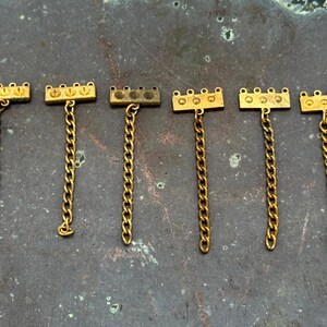 May include: Six gold-toned metal jewelry pieces with a rectangular top and a chain. The rectangular top has three or four small circular details. The chains are a linked design. The pieces are displayed on a mottled, dark surface.
