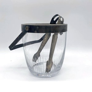 Peut inclure: Un seau à glace en verre transparent avec un bord et une poignée en métal argenté. Le seau présente des lignes verticales gravées et comprend une paire de pinces à glace. Les pinces et la poignée sont en métal foncé.