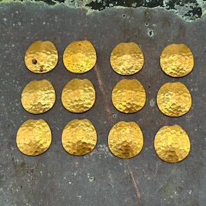 May include: Twelve hammered gold-colored metal oval shapes with a textured surface are arranged on a dark, mottled background. Each piece has a small hole at the top, suggesting they are components for jewelry or crafts.