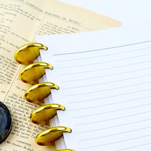 May include: A notebook with lined pages and gold-colored disc-bound rings. A vintage paper with French text and a small, round, dark blue container with a gold emblem are also present.