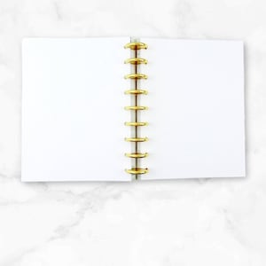 May include: Open white notebook with gold spiral binding. The notebook is lying open on a white marble surface. The pages are blank and ready for notes or sketches. The gold binding adds a touch of elegance.