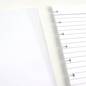 May include: Two stacks of white paper with pre-punched holes for a binder. One stack has a dot grid pattern, and the other has horizontal lines. The pages are clean and ready for notes or planning.