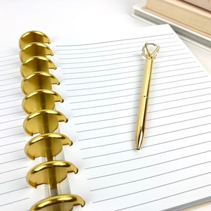 May include: A gold pen with a diamond-shaped crystal top rests on a lined notebook. The notebook is open, revealing the pages and the gold disc-shaped binding. The pen and binding have a shiny, metallic finish. A stack of books is in the background.