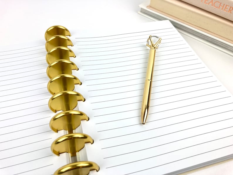 May include: A gold pen with a diamond-shaped crystal top rests on a lined notebook. The notebook is open, revealing the pages and the gold disc-shaped binding. The pen and binding have a shiny, metallic finish. A stack of books is in the background.