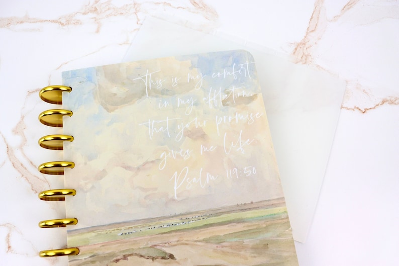 May include: A spiral-bound planner with a landscape design and gold-colored metal rings. The cover features a watercolor painting of a field and sky, with the text "This is my comfort in my affliction that your promise gives me life Psalm 119:50."