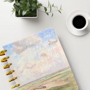 May include: A spiral-bound notebook with a watercolor design featuring a sky and landscape scene. The cover has gold-colored discs and white text that reads "This is my comfort in my affliction that your promise gives me life Psalm 119:50."