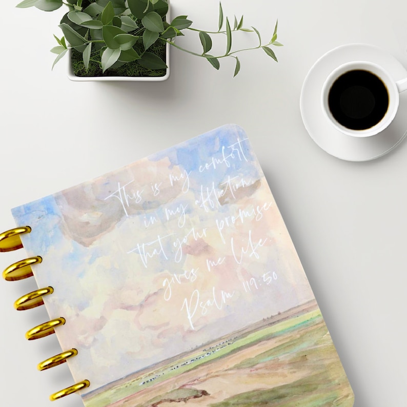 May include: A spiral-bound notebook with a watercolor design featuring a sky and landscape scene. The cover has gold-colored discs and white text that reads "This is my comfort in my affliction that your promise gives me life Psalm 119:50."