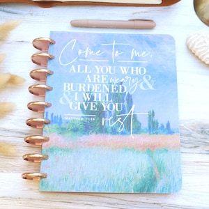 May include: A disc-bound planner featuring a landscape painting cover with the white text: Come to me, ALL YOU WHO ARE WEARY & BURDENED & I WILL GIVE YOU REST, MATTHEW 11:28. The planner has metallic copper-colored discs along the left side.