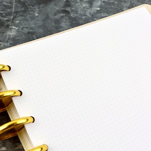 May include: A close-up of a white dot grid notebook with gold-colored metal rings. The notebook is open, revealing blank pages. The background is a dark gray, textured surface.