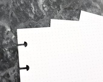 May include: Close-up of white dot grid paper with two punched holes for a binder. The paper is on a dark gray surface. The paper has a subtle dot pattern, perfect for journaling or note-taking.
