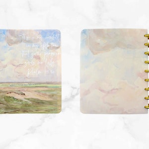 May include: A spiral-bound notebook with a landscape painting on the cover, featuring a field, sky, and clouds. The cover has gold-colored rings. Text on the cover reads: "This is my comfort in my affliction that your promise gives me life. Psalm 119:50."