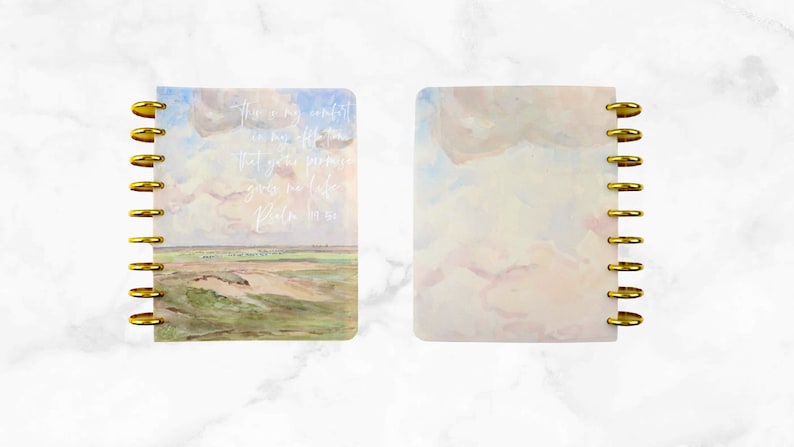 May include: A spiral-bound notebook with a landscape painting on the cover, featuring a field, sky, and clouds. The cover has gold-colored rings. Text on the cover reads: "This is my comfort in my affliction that your promise gives me life. Psalm 119:50."
