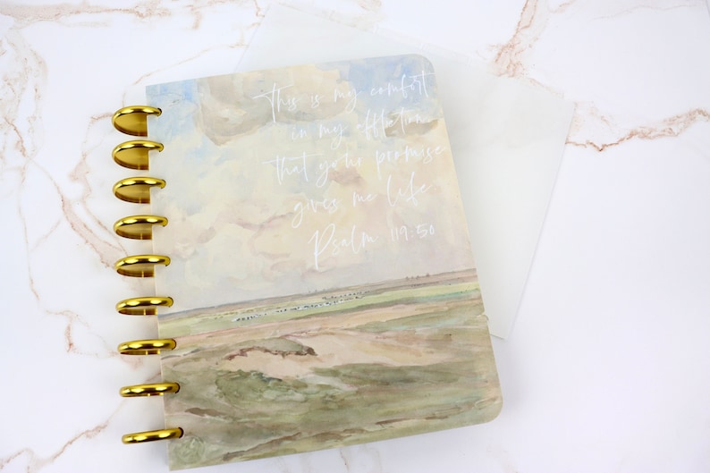 May include: A discbound notebook with a landscape watercolor design and gold metal rings. The cover features a sky and field scene with the text "This is my comfort in my affliction that your promise gives me life. Psalm 119:50."