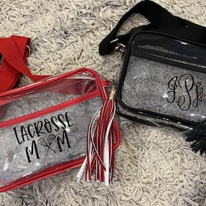 May include: Two clear plastic crossbody bags with red and black straps and tassels. One bag has a black and white lacrosse stick design with the text "Lacrosse Mom". The other bag has a black monogram.