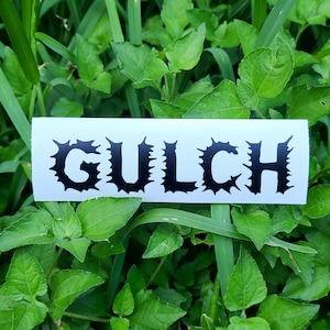 May include: A white sticker with black text that reads "GULCH" in a jagged, spiky font. The sticker is placed on a bed of green grass.