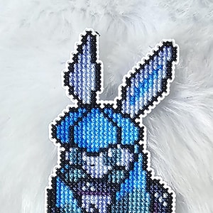 Peut inclure: Marque-page fait à la main avec un motif de point de croix bleu et blanc représentant une créature ressemblant à un lapin. Le marque-page est rectangulaire avec une bordure blanche et une boule rouge et blanche en bas.