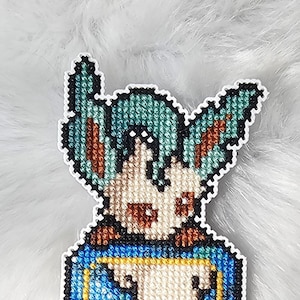 Peut inclure: Marque-page fait à la main en point de croix représentant un personnage de dessin animé aux cheveux turquoise et au corps crème. Le marque-page a une bordure bleue avec un contour jaune et un design Pokeball en bas. Le fond est blanc.