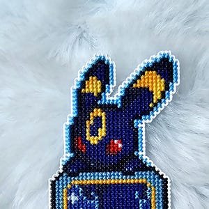 Peut inclure: Marque-page en point de croix représentant Umbreon bleu foncé avec des accents jaunes. Le marque-page est de forme rectangulaire avec une scène intérieure détaillée dans les tons violets, bleus et blancs, et une bordure dorée. Posé sur une surface blanche et duveteuse.