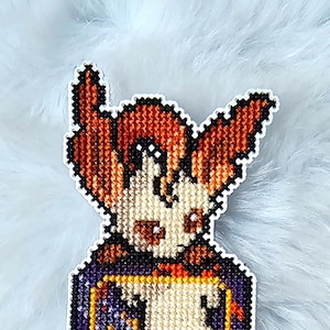 Peut inclure: Marque-page en point de croix pixelisé représentant un personnage aux cheveux roux dans un cadre violet et doré. Le motif comprend une silhouette stylisée avec une queue rouge et un petit emblème rond en bas. Le fond est blanc.