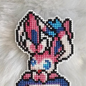 Peut inclure: Un marque-page en point de croix rose et bleu représentant un personnage de Pokemon, avec un fond de galaxie et un Poké Ball en bas.