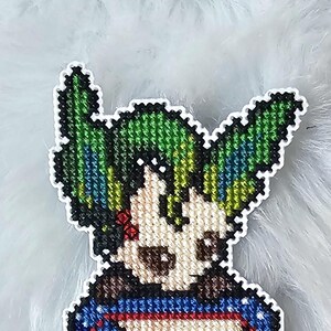 Peut inclure: Marque-page fait à la main en point de croix représentant un personnage fantaisiste aux cheveux verts et un fond bleu et blanc. Le marque-page a une bordure rouge et un petit motif de Pokéball en bas. Le personnage a la peau claire et sourit.