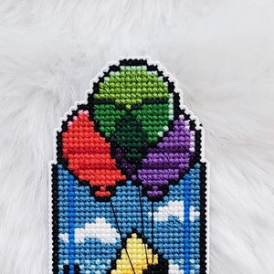 Może przedstawiać: Zakładka haftowana krzyżykiem przedstawiająca żółtego Pikachua trzymającego kolorowe balony. Tło jest niebieskie z białymi chmurami. Zakładka jest oprawiona w białą ramkę i zawiera tekst "Melissassy Stitchery".