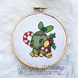 May include: A cross-stitch pattern of a green and brown Pokemon character holding a candy cane. The character is wearing a red and white candy cane and has a holly sprig on its head. The pattern is framed in a wooden hoop.