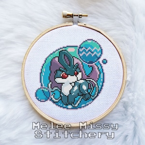 Könnte beinhalten: Ein Kreuzstich-Stickrahmen mit einem Pixelbild eines blauen und violetten Wesens mit roten Augen. Das Wesen ist von blauen und weißen Blasen umgeben. Der Rahmen ist aus Holz und hat einen weißen Hintergrund. Der Text "Melee Missy Stitchery" ist unter dem Rahmen gedruckt.