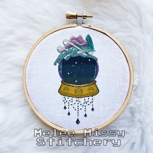 Könnte beinhalten: Ein Kreuzstichmuster eines Schneekugels mit einem blauen und violetten Wesen darin. Der Schneekugel steht auf einem goldenen Sockel mit einer Perlenkette, die daran hängt. Der Hintergrund ist weiß.