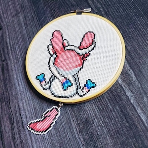 May include: A handmade cross-stitch embroidery in a wooden hoop. The design features a pink and white cartoon character with blue accents. A small, matching charm hangs below. The artwork is displayed on a dark wood surface.