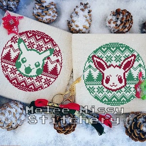 May include: Two cross-stitch patterns featuring a white Pokemon character with red and green details. The patterns are on a beige canvas and are surrounded by pine cones and embroidery floss.