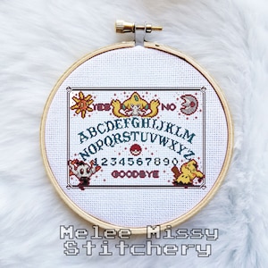Könnte beinhalten: Ein weißer Stickrahmen mit einem Kreuzstich-Design eines Pokemon Ouija-Bretts. Das Brett zeigt die Buchstaben A-Z, die Zahlen 1-9 und 0, die Wörter "YES", "NO" und "GOODBYE". Drei Pokemon-Charaktere sind auf dem Brett abgebildet: ein gelber Pikachu, ein braunes und rotes Gastly und ein gelbes und weißes Sylveon.