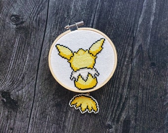 May include: A yellow and white embroidered cross-stitch of a Pokemon character, Eevee, in a wooden hoop.