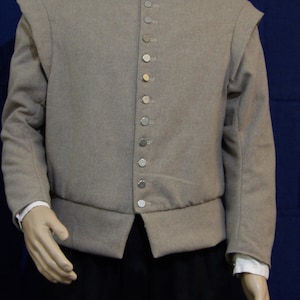 May include: A tan-colored historical jacket with a high collar and a row of silver-colored buttons down the front. The jacket has long sleeves and a tailored fit, with a slight flare at the waist. The jacket is part of a historical costume.