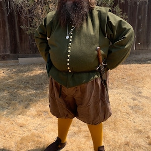 Op de afbeelding: Een persoon draagt een groene tuniek met gouden knopen, een bruine korte broek en gele kousen. De outfit omvat een bruine hoed met veren en bruine leren schoenen. Een zwaard is aan de zijkant bevestigd.
