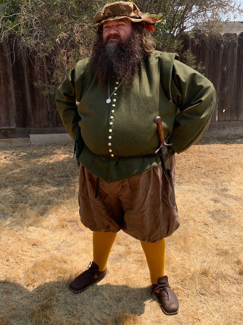 Op de afbeelding: Een persoon draagt een groene tuniek met gouden knopen, een bruine korte broek en gele kousen. De outfit omvat een bruine hoed met veren en bruine leren schoenen. Een zwaard is aan de zijkant bevestigd.